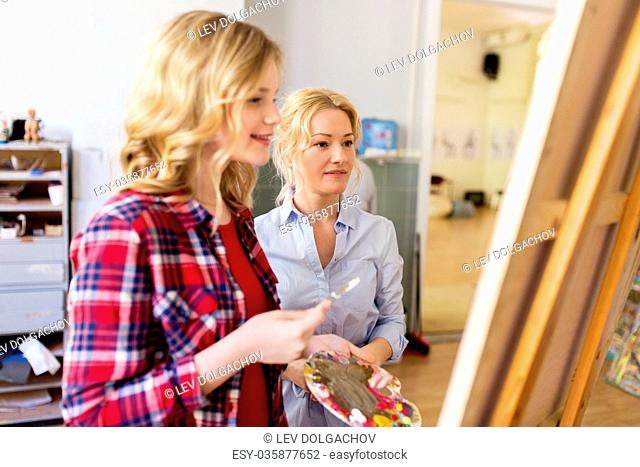 creativity, education and people concept - artists or student girl with palette and painting knife and teacher discussing picture on easel at art school studio