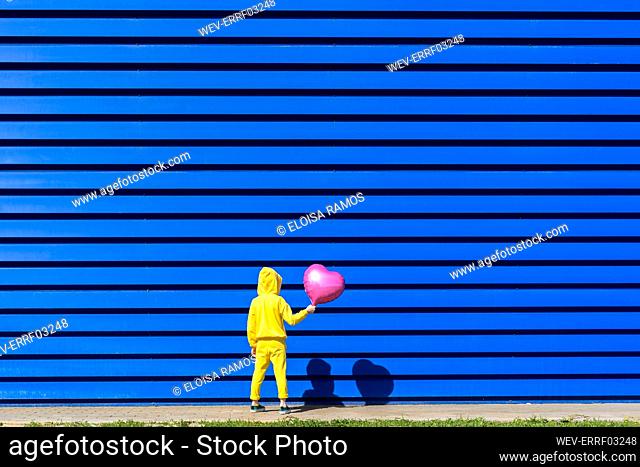 Back view of little girl with pink balloon wearing yellow tracksuit standing in front of blue background