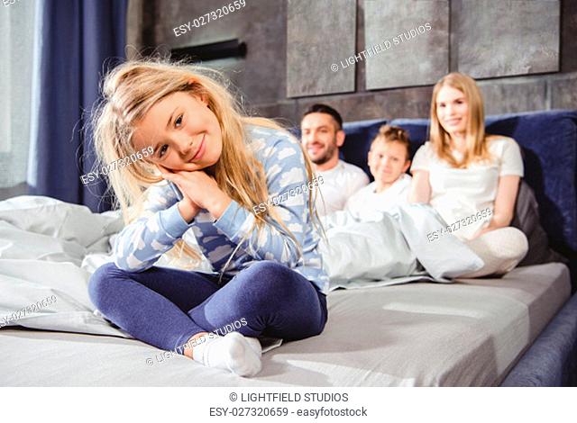 Happy little girl sitting on bed with her family and looking at camera