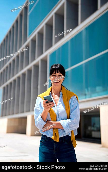 Happy businesswoman holding smart phone standing in front of building
