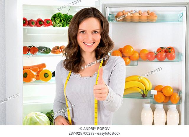 Cheerful Woman With Measuring Tape Showing Thumbs Up Sign In Front Of Open Refrigerator