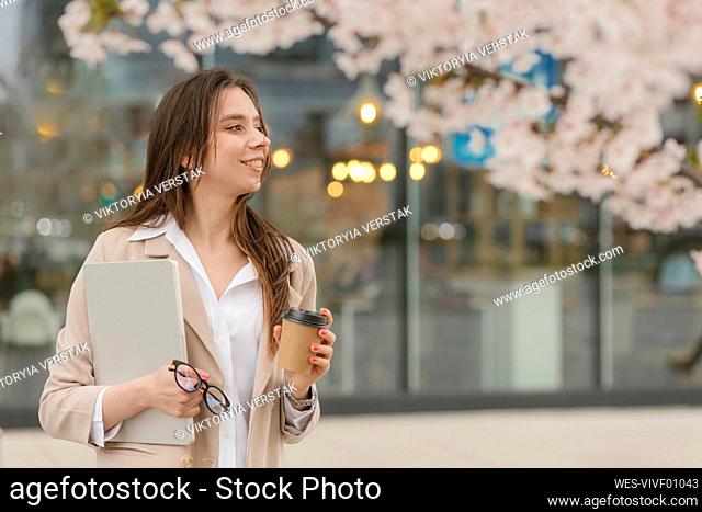 Happy businesswoman standing with coffee cup and laptop