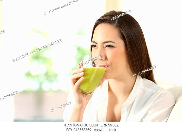 Happy woman drinking a green vegetable juice sitting on a couch in the living room at home
