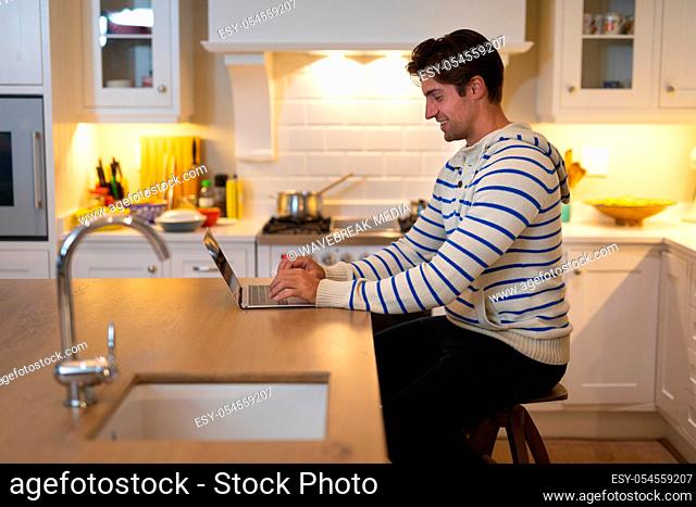 Side view of a Caucasian man at home wearing a blue striped sweater, sitting in the kitchen at the kitchen island, using a laptop computer and smiling