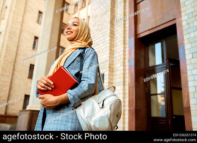 Arab girl with books poses at university entrance. Muslim woman in hijab holds textbooks outdoors. Religion and education