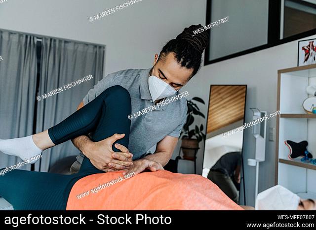 Physiotherapist in protective face mask doing therapy on knee of female patient in medical practice during pandemic