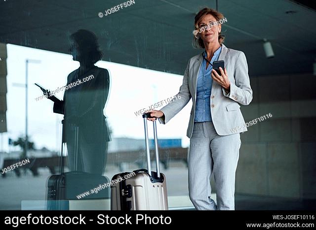 Smiling businesswoman holding mobile phone standing with suitcase in front of glass wall