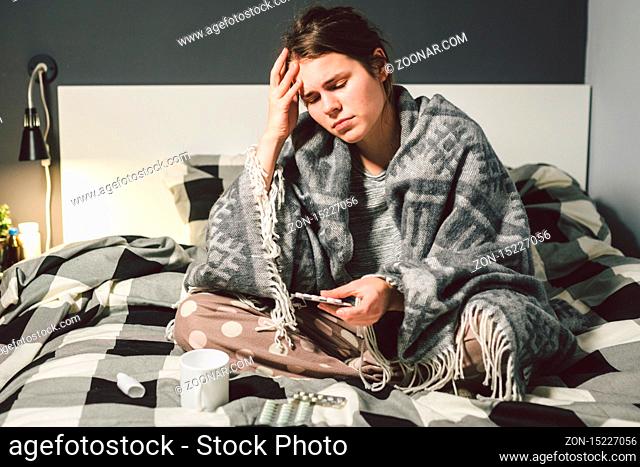 Subject disease and treatment. young Caucasian woman sits home bedroom bed wrapped in body blanket with high fever, fever holds strong headache behind head