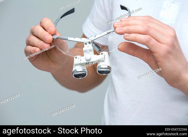 Doctor in white t-shirt holding the dental binocular loupes in his hands. Studio photo on the gray background. Horizontal