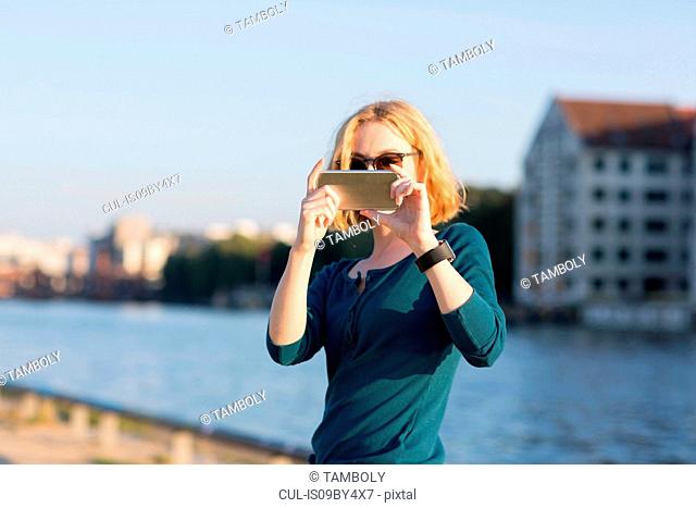 Young woman taking photograph with smartphone by river in summer, Berlin, Germany