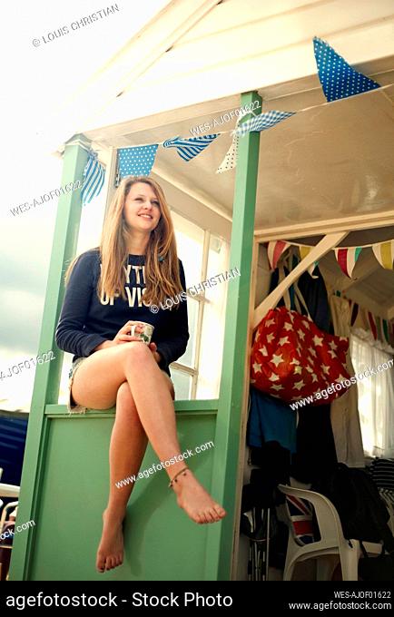 Smiling teenage girl looking away while sitting on window of beach hut