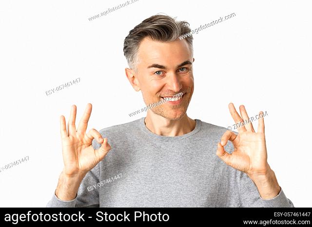 Close-up of handsome middle-aged man, smiling and showing okay signs, approve something or recommending, standing over white background
