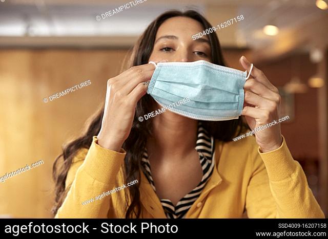Portrait of young ethnic woman putting on face mask wearing yellow sweater with black and white striped blouse sitting at bar in kitchen of downtown loft