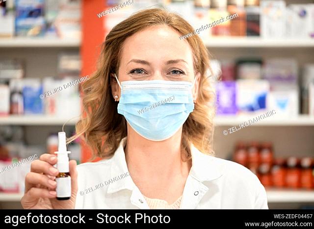 Female pharmacist wearing protective face mask holding bottle of nasal spray in pharmacy store