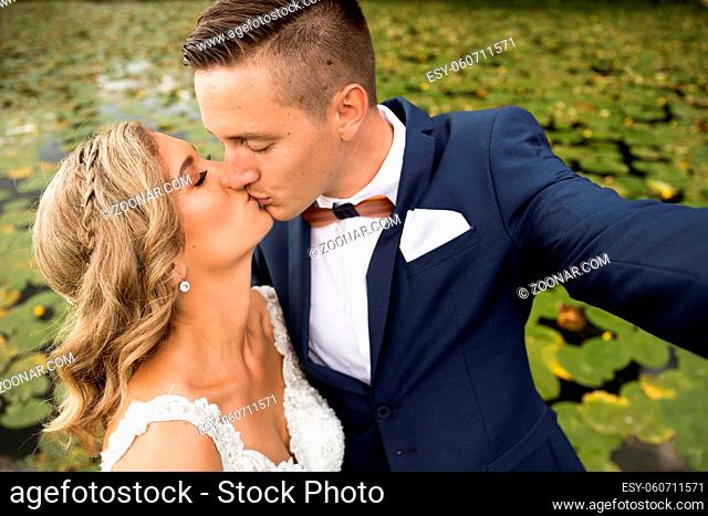 Wedding selfie. Bride and groom kissing passionately outdoors in the park. Caucasian happy romantic young couple celebrating their marriage