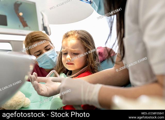 Dentist talking with cute little girl sitting by mother at medical clinic