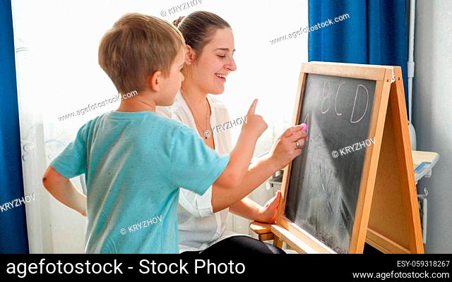 Happy mother with smiling son learning and writing alpabet on blackboard with chalks at home