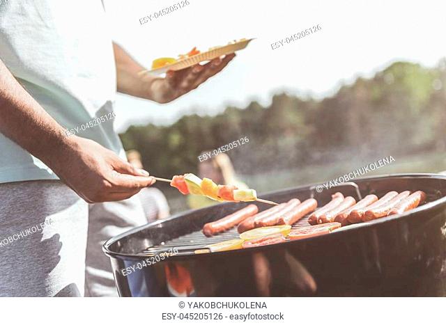 male hands holding vegetable skewers and plate while standing near grill. Vegies and sausages are on grate. Close up