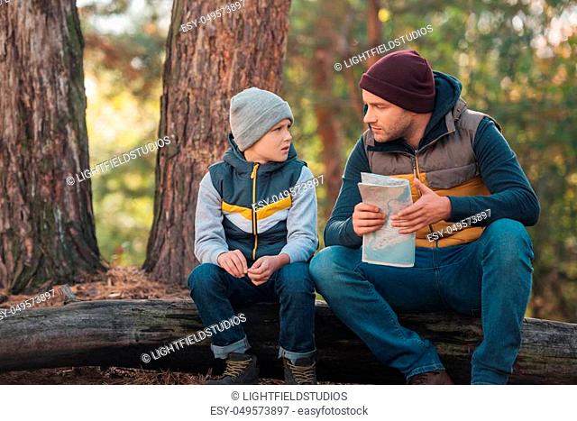 cute little boy looking at father holding map while sitting on log in forest