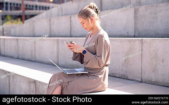 Side view of trendy young woman with laptop sitting on concrete steps and browsing social media on laptop on summer day in city