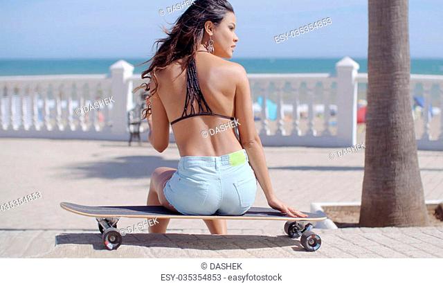 Shapely young woman sitting waiting on a skateboard overlooking the ocean and beach on a seafront promenade looking to the right as she waits for someone view...