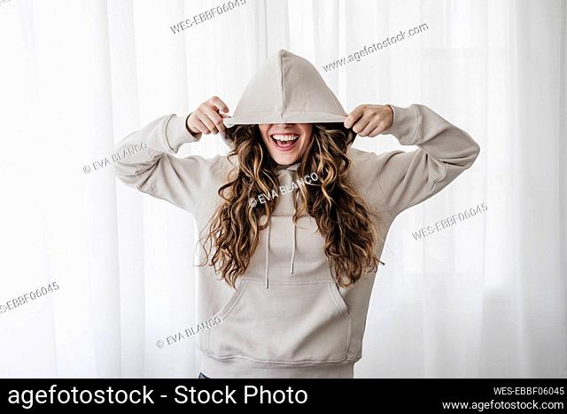 Playful woman covering eyes with hood at home