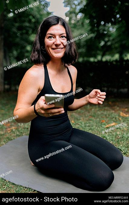 Smiling yoga teacher with mobile phone sitting on exercise mat in park