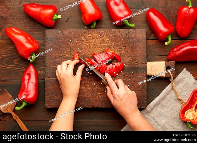 woman cuts red bell pepper into pieces on wooden board, brown wooden table, top view
