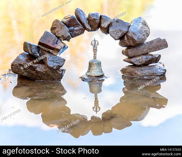 rocks piled up in a half circle reflected in water looking like a zero with a bell in the middle