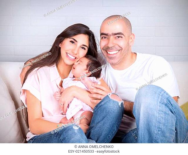 Portrait of happy cheerful family sitting on the couch at home, young parents enjoying time spent with their adorable newborn daughter