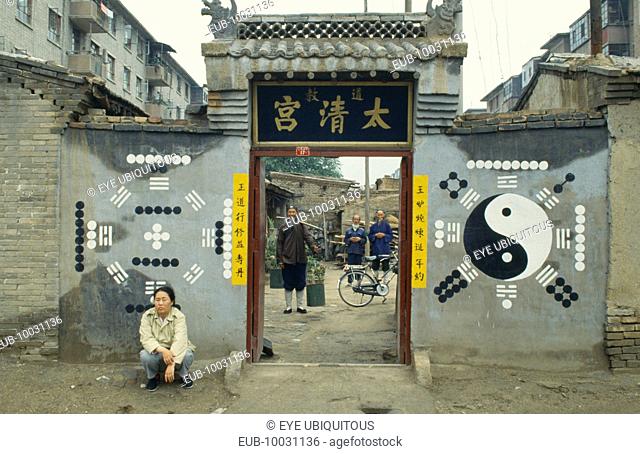 Monks or priests seen through the entrance of Daoist temple with trigram and yin yang symbols on the exterior walls and person crouched outside