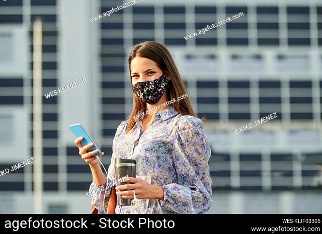 Young woman wearing face mask holding thermos while using mobile phone in city
