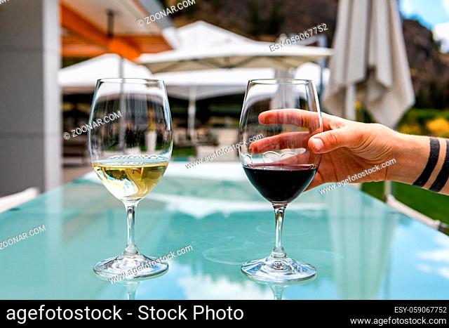 hand on red wine glass, Two glasses of red and white wine, pair of red pinot noir and white chardonnay on a glass table in beautiful patio outdoors
