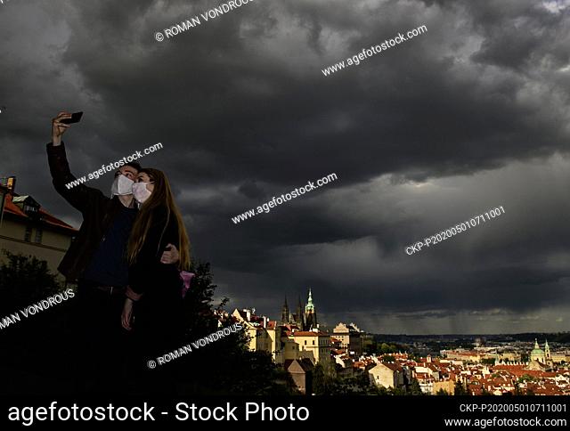 A young couple wearing face masks takes a selfie with heavy clouds above Prague centre in background in park near Strahov Monastery in Prague, Czech Republic