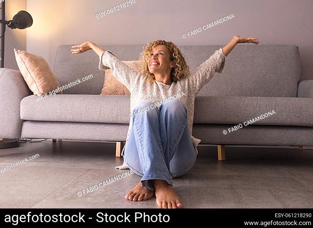 Cheerful young woman sitting on floor with arms raised at home. Thoughtful caucasian woman smiling sitting in living room