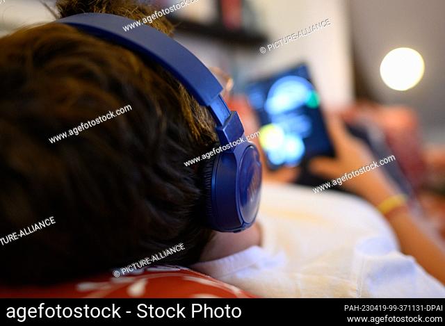 PRODUCTION - 18 April 2023, Saxony, Dresden: ILLUSTRATION - A woman is lying on a sofa with headphones on, holding an iPad tablet with an open app for audio...