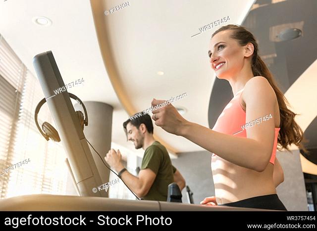 Happy young woman listening to music while running beside a handsome man on a modern treadmill with touch screen and headphones