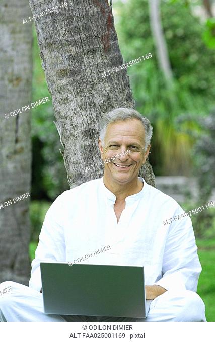 Mature man sitting, leaning against tree, using laptop computer, smiling at camera