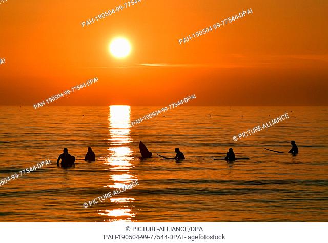 21 April 2019, Denmark, Klitmöller: Surfers wait for a wave in the water of the North Sea, while the colourful sunset bathes the sky and the sea in a warm light