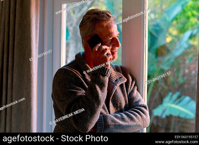 Happy caucasian senior man standing at window and making call