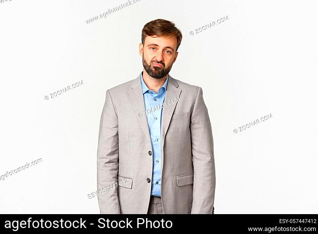 Portrait of skeptical and unamused bearded male employee in grey suit, looking disappointed and sad, standing over white background