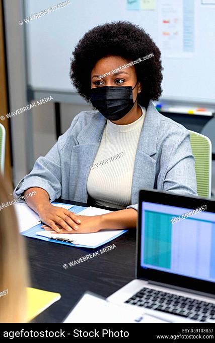 Portrait of african american businesswoman wearing face mask sitting at desk in meeting room