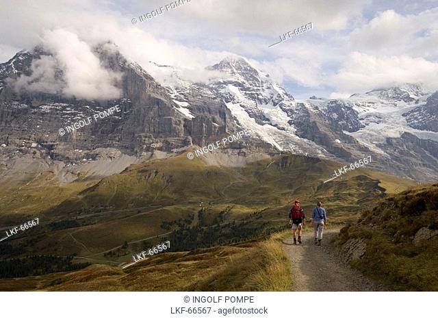 Couple hiking along Maennlichen Panoramaweg panorama hiking path, view to Kleine Scheidegg 2061 m, Eiger 3970 m, Moench 4107 m and Jungfrau 4158 m in background