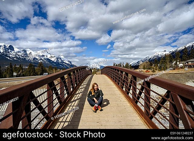 A middle aged woman sitting in the middle of a wood pedestrian bridge over Cougar Creek in Canmore, Canada with snow capped mountain peaks in the background