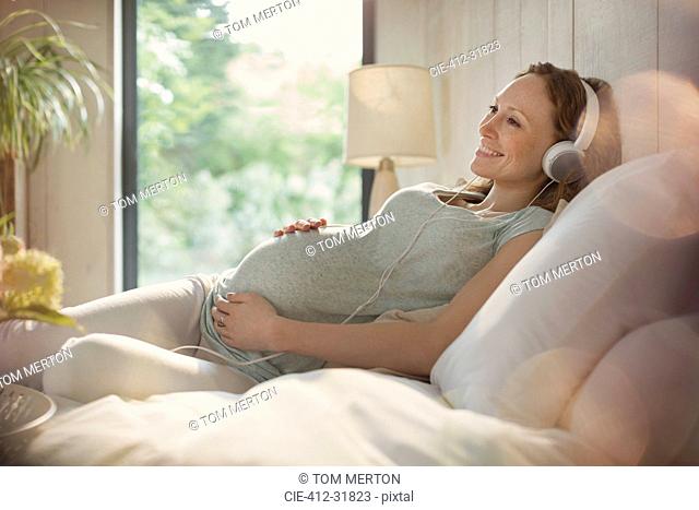 Smiling pregnant woman relaxing listening to music with headphones in bed