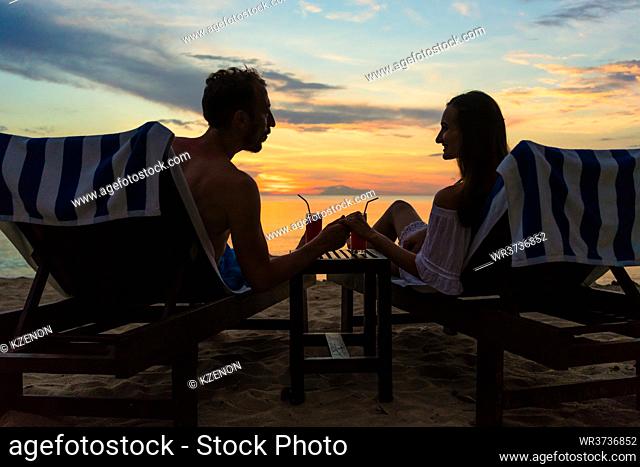 Rear view of a young romantic couple in love, sitting on wooden chairs while drinking cocktails on a tropical beach at sunset during vacation or honeymoon in...