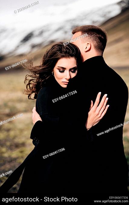 Wedding couple on a background of snowy mountains. The bride and groom in black coats are hugging in a field of moss and yellow grass
