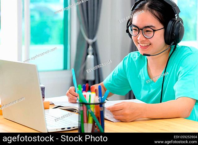 Asian young woman student with glasses headphones girl study happy writing note on a book looking video conference laptop computer university class online...