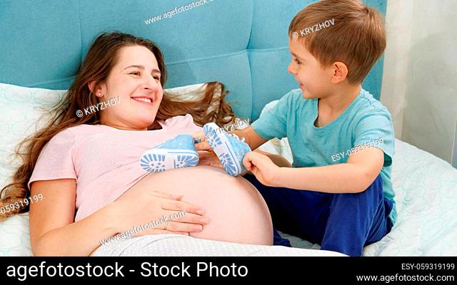 Funny shot of little boy playing and dancing with newborn baby boots on big belly of pregnant mother.Concept of pregnancy, preparing and expecting child