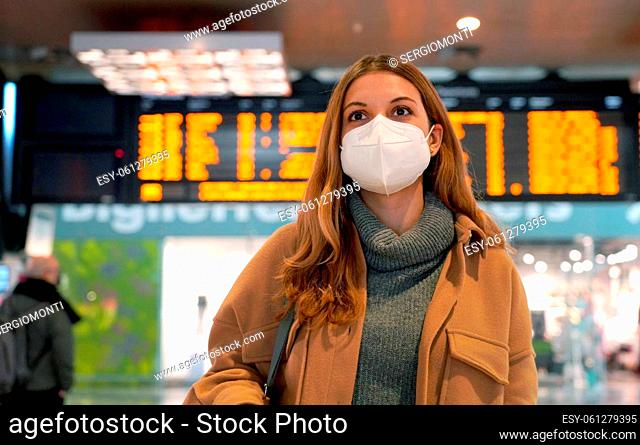 Business woman wearing face mask FFP2 KN95 at train station and keeping social distance. Young woman waiting for the train with timetables on background